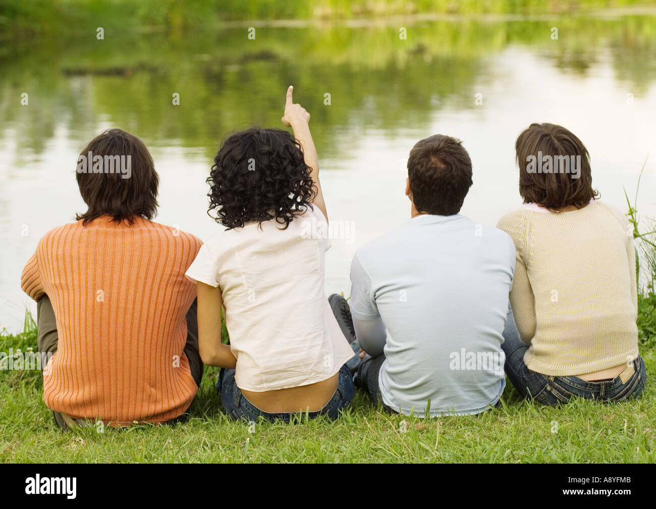 Four friends sitting near the edge of a pond, one pointing, rear ...
