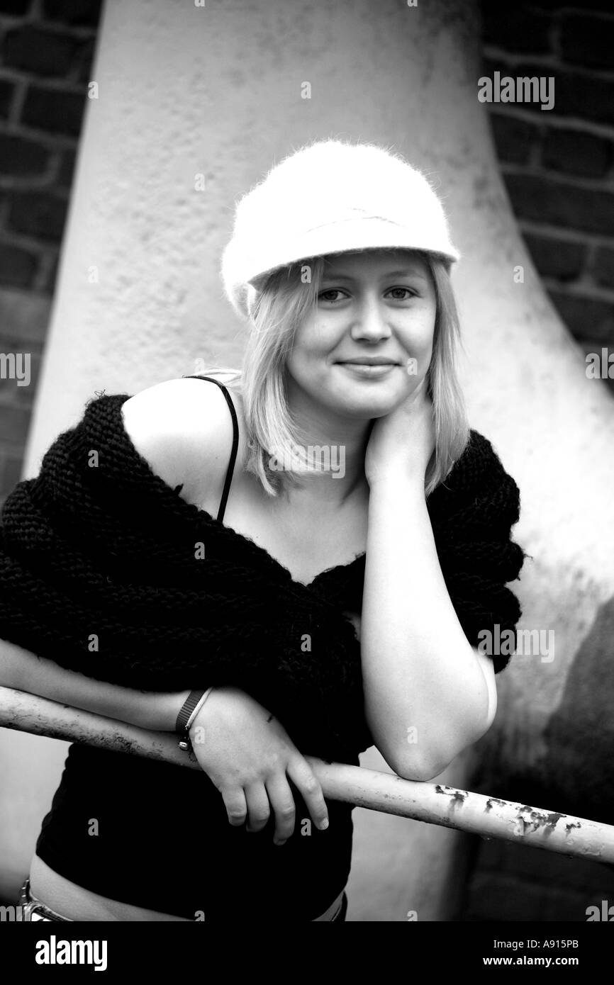 Vertical black and white portrait of young woman in fluffy hat, leaning on railings outdoors in derelict location smiling Stock Photo