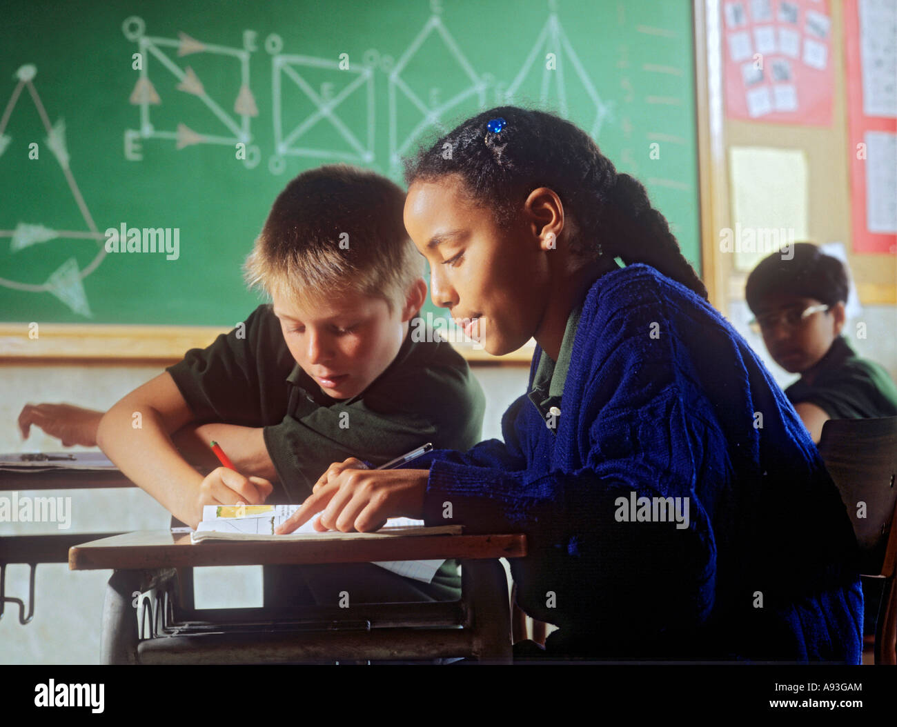 Junior schoolgirl in classroom working with other pupils, geometry blackboard behind Stock Photo