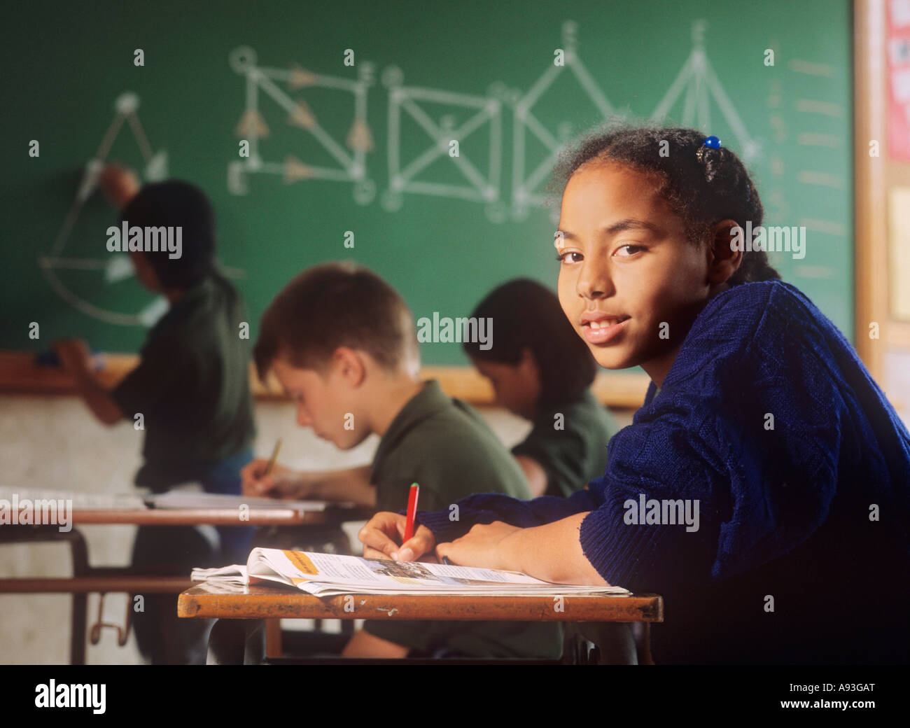 Junior schoolgirl in classroom with other pupils and geometry blackboard behind Stock Photo