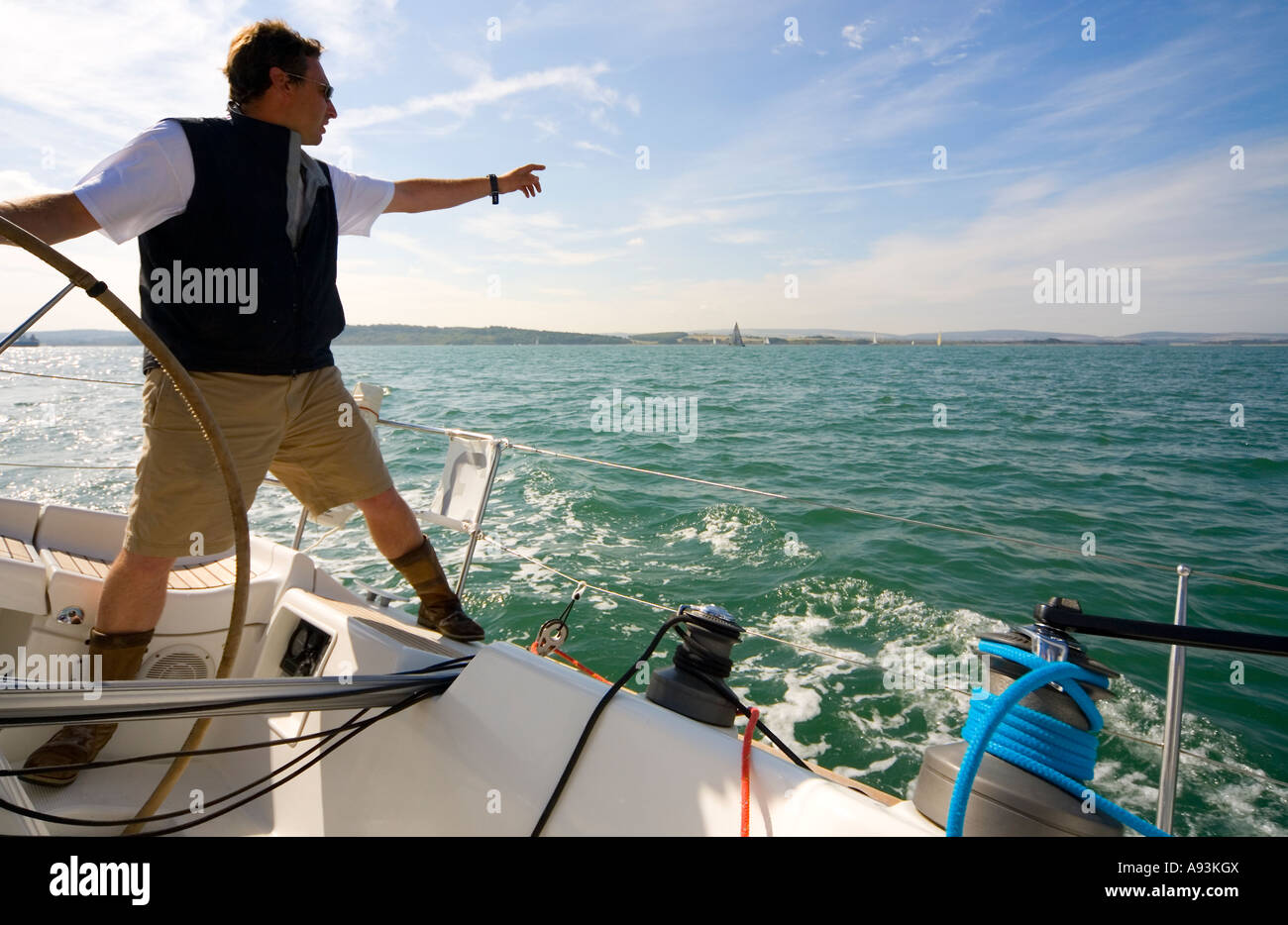 The skipper of a yacht points towards the horizon while keeping one hand on the boat's wheel Stock Photo