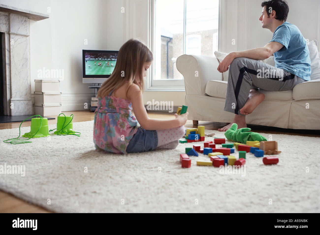 Girl (5-6) sitting on floor, playing with blocks, father watching television Stock Photo