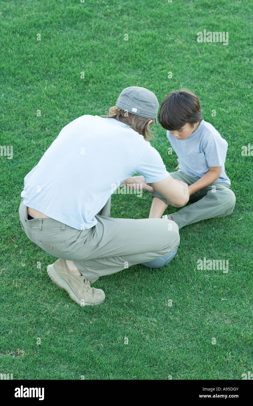 Boy sitting on grass with pants leg rolled up, man leaning over him