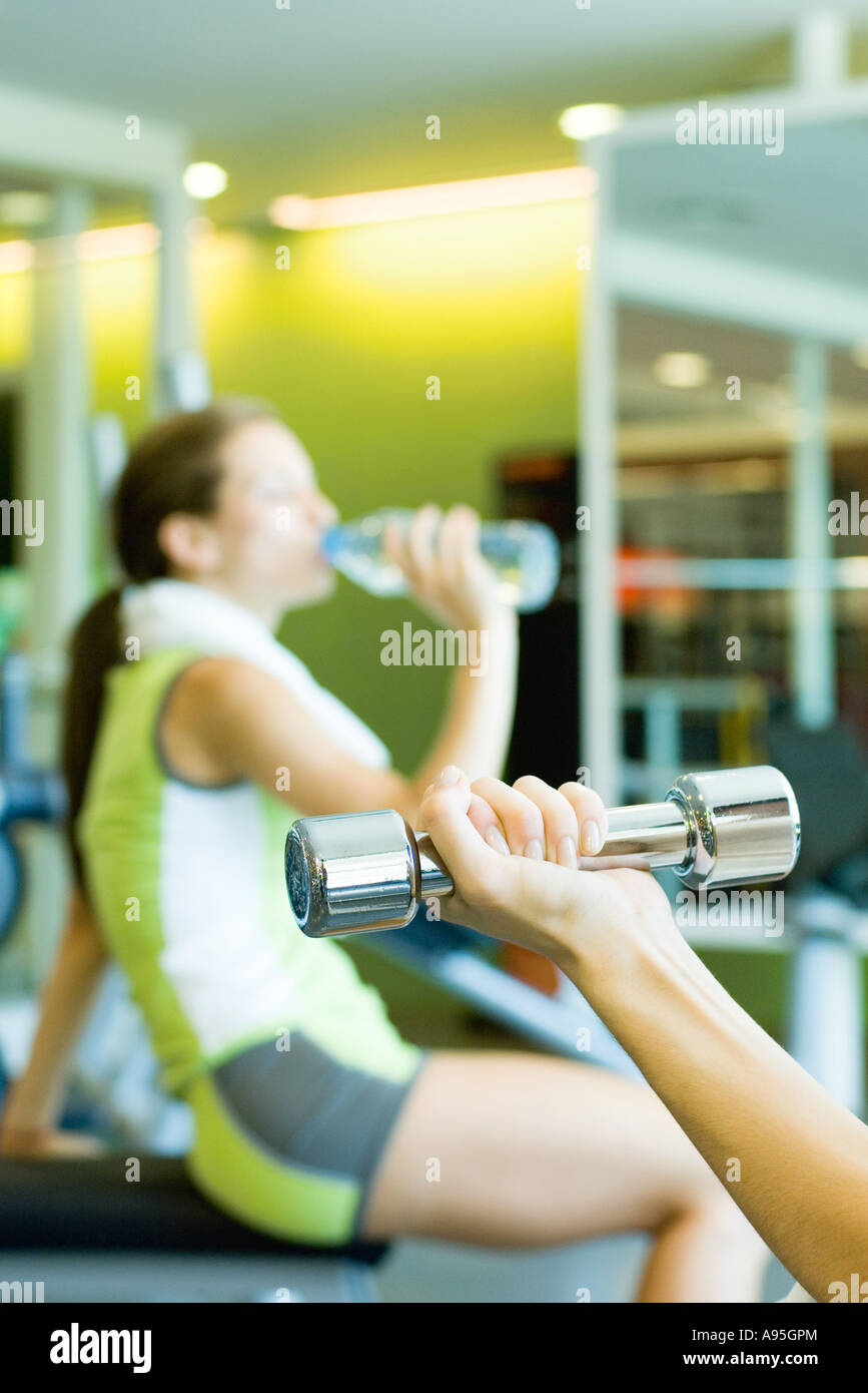 Woman drinking bottle of water, focus on second woman's arm holding dumbbell in foreground Stock Photo