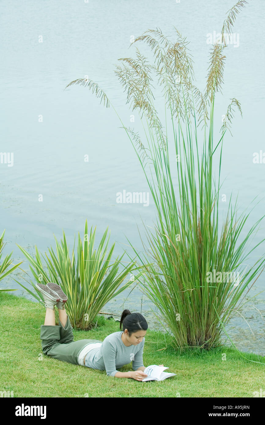 Young woman lying on grass by edge of lake, reading Stock Photo