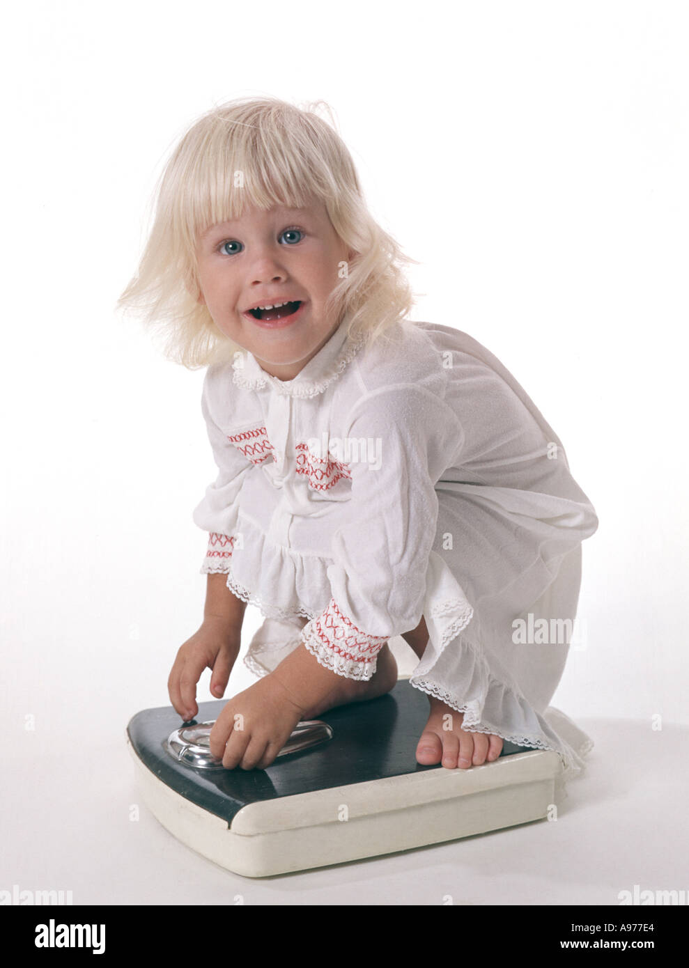 small girl in nightgown squatting on bathroom scales Stock Photo