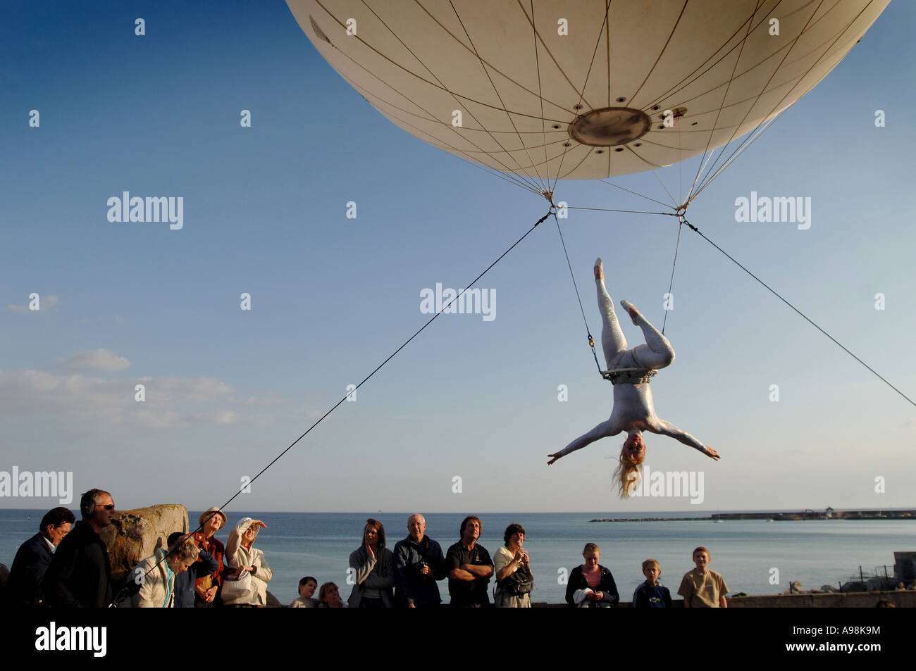 A giant helium balloon with an aerial trapeze artist suspended beneath drifted above spectators on Dorset s Jurassic Coast Arts Stock Photo
