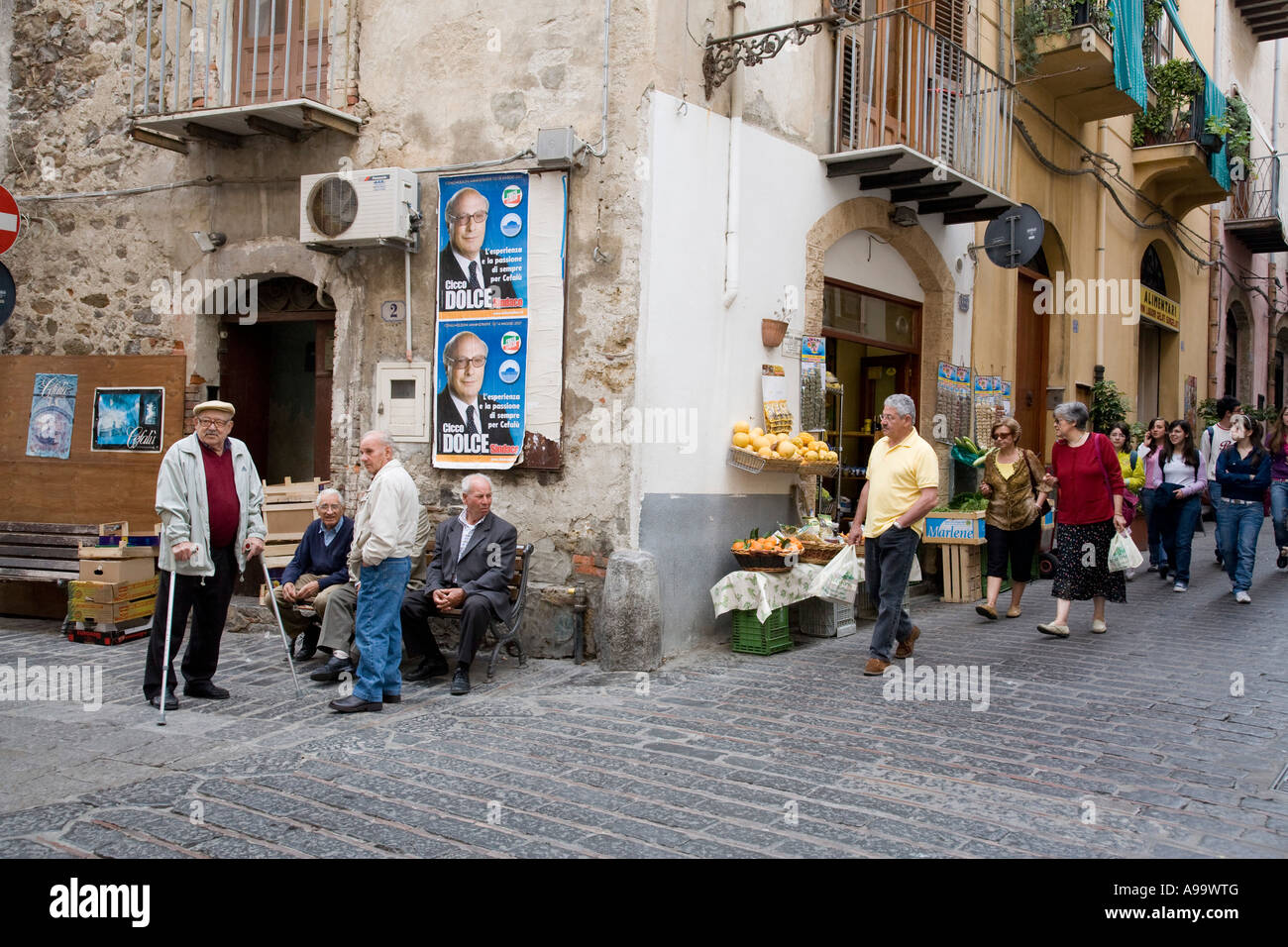 Sicilian men sitting on bench under a political poster in front of alimentari Corso Ruggero Cefalu Sicily Italy Stock Photo