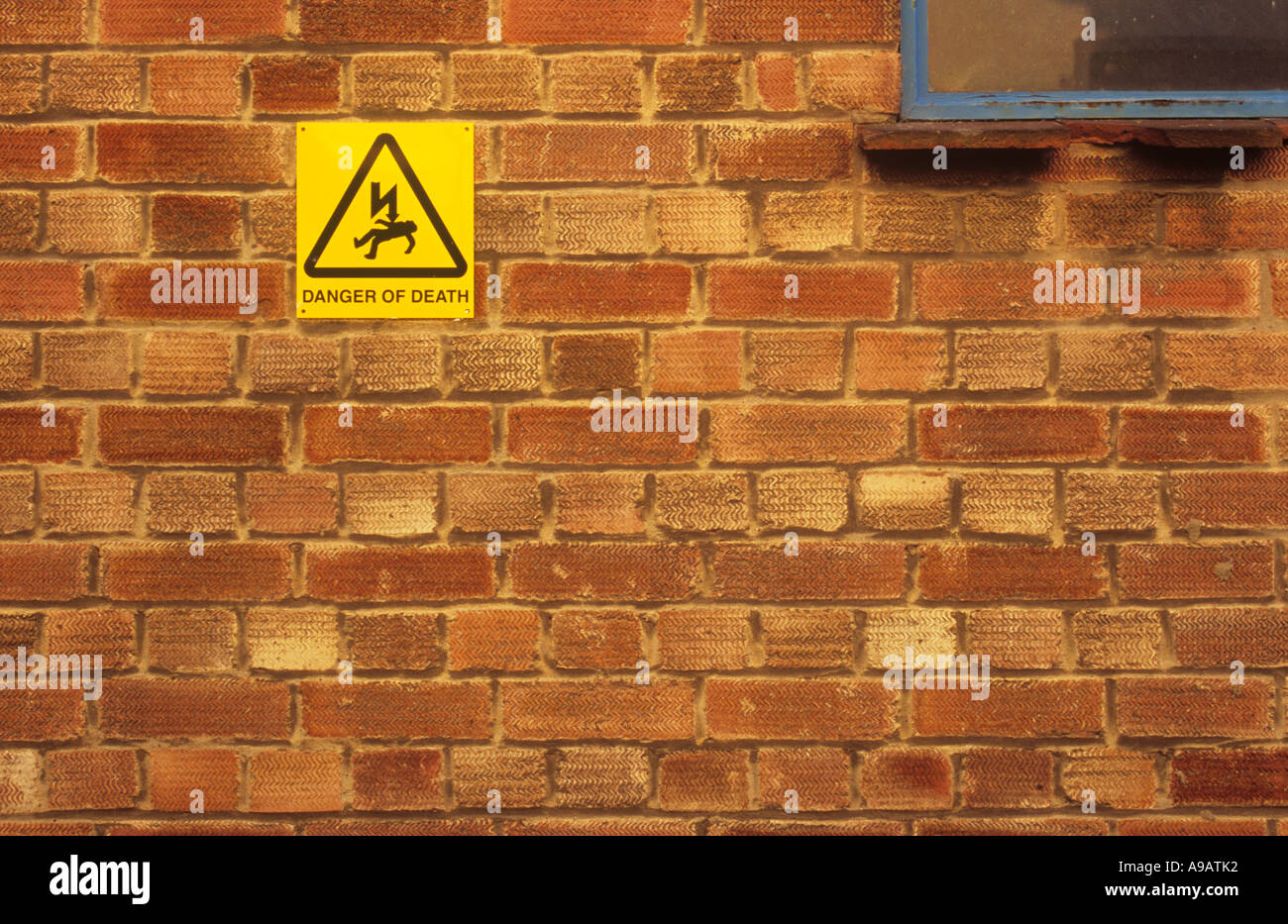Orange brickwall with rusting blue window frame and yellow sign with iconic man being electrocuted and words Danger of death Stock Photo