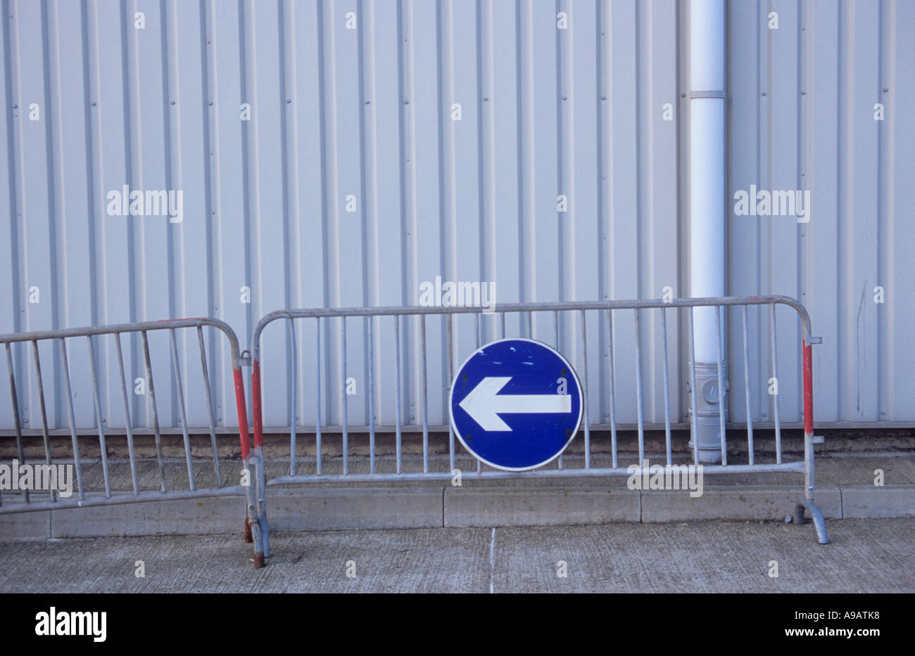 Traffic barrier or mobile railing with blue and white Turn or Keep left sign in front of pale grey slatted warehouse walls Stock Photo