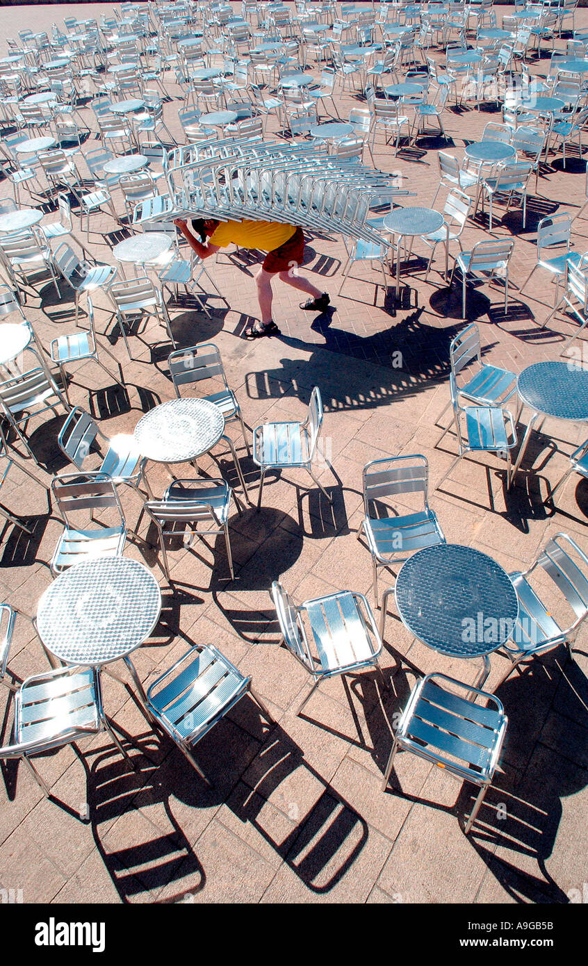 A young man hauls a stack of 16 metal chairs across the terrace in front of Gemini Lounge and Beach Bar on Brighton  seafront Stock Photo