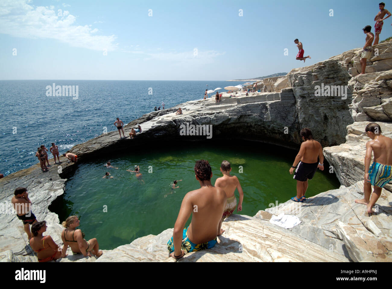 Holidaymakers sitting on the rocks around a natural circular pool beside the sea watching a boy jumping in. Stock Photo