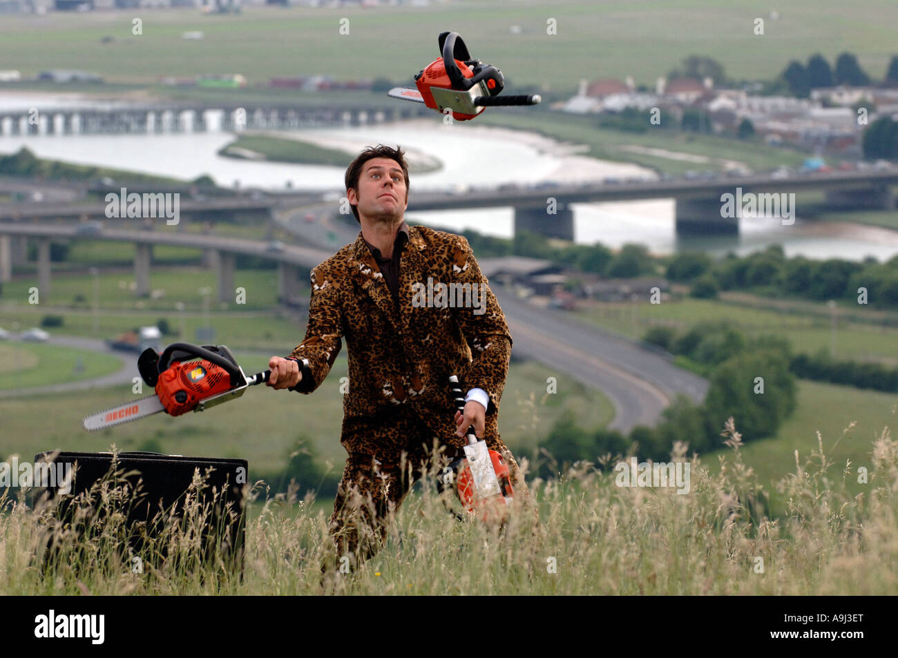 Young entertainer in leopardskin jacket juggling three orange chainsaws on the South Downs Stock Photo