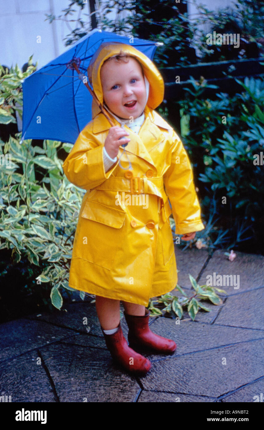 Small child in a bright yellow raincoat and sou'wester hat, holding a blue umbrella, in red wellie boots, in a UK garden. Circa 1960s Stock Photo