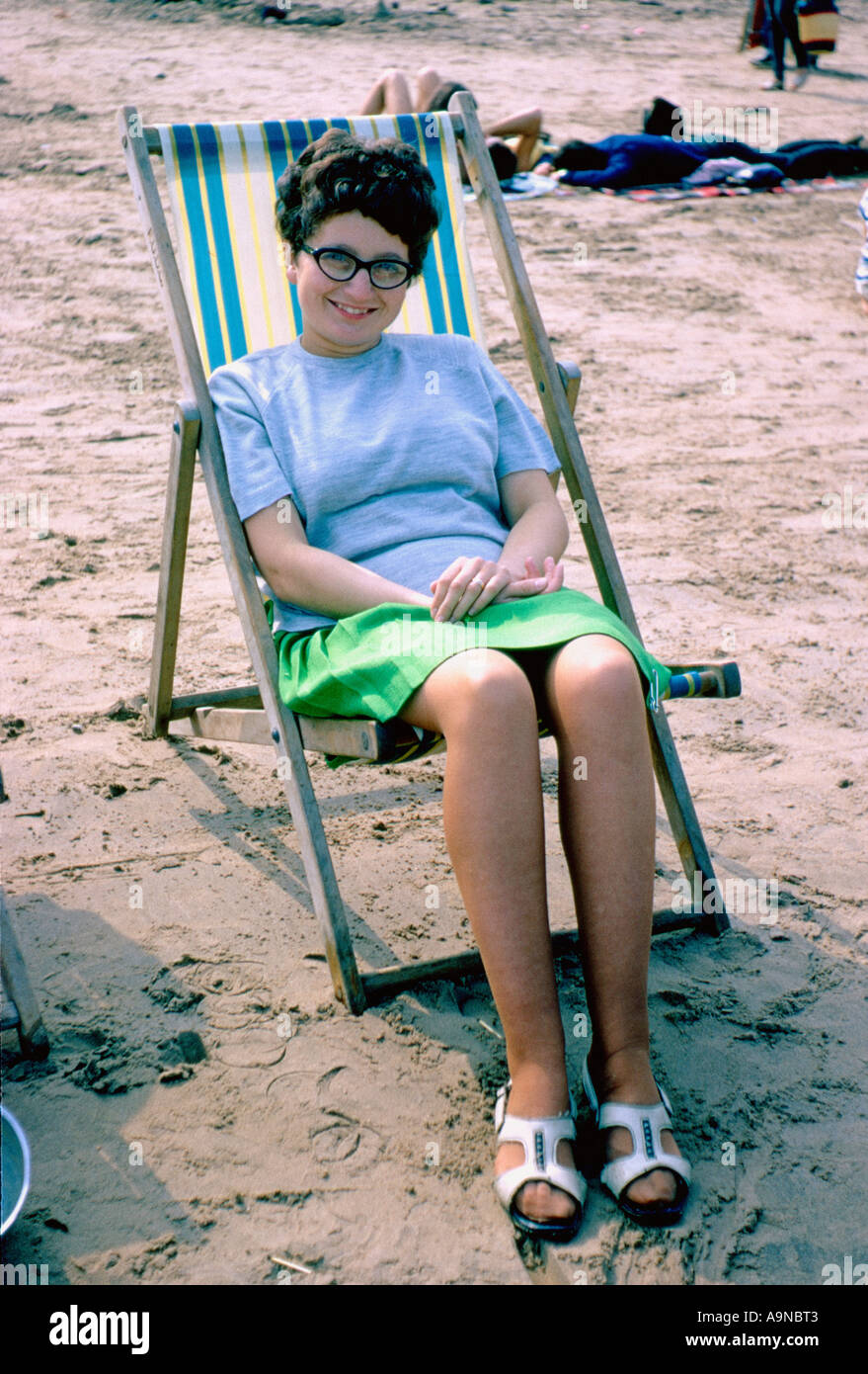1960s: Young Caucasian woman in a mini skirt and cat-eye glasses, relaxing in a deckchair on a UK beach Stock Photo