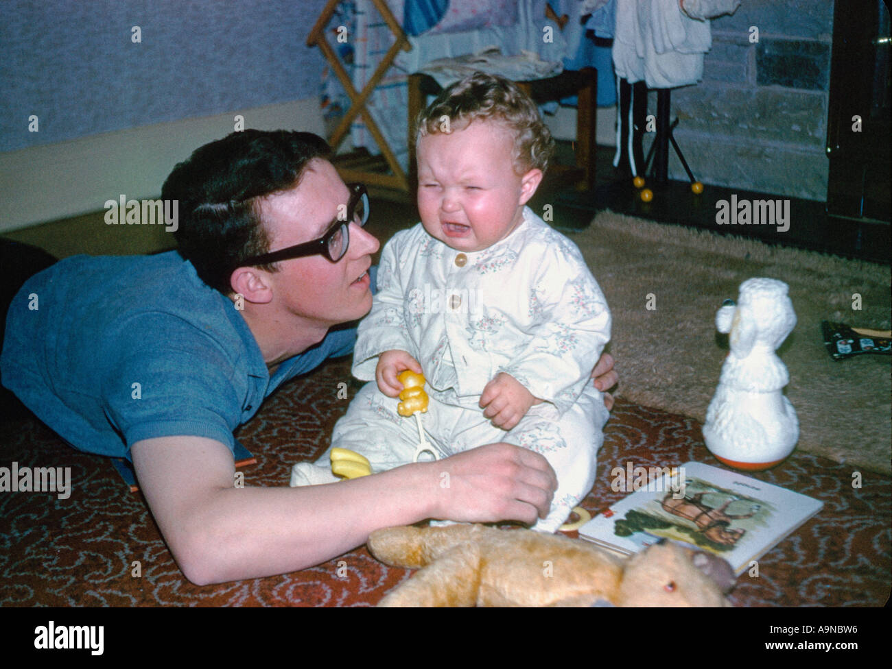 Young caucasain father laid on the floor next to a crying baby sitting on the floor in a typical 1960s home. UK. Archive image Stock Photo
