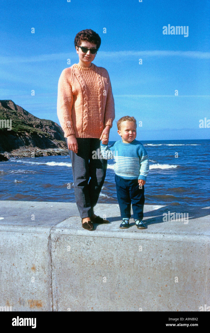 Young Caucasian woman and small child in knitted jumpers, standing hand-in-hand on a UK seafront wall, 1960s Stock Photo