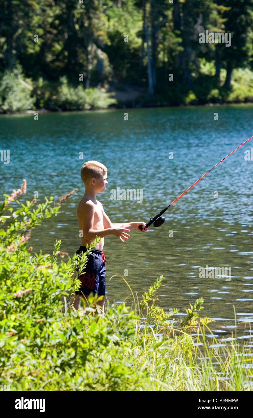 Image of a young boy fishing from the bank of a lake He s dressed for summer in baggy shorts and no shirt Stock Photo