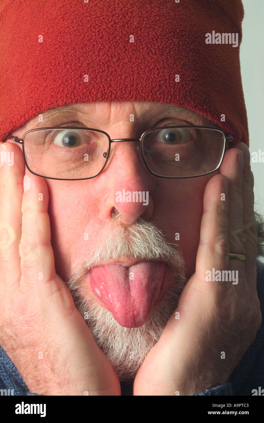 Elderly man in warm red hat with hands to face poking out tongue Stock Photo