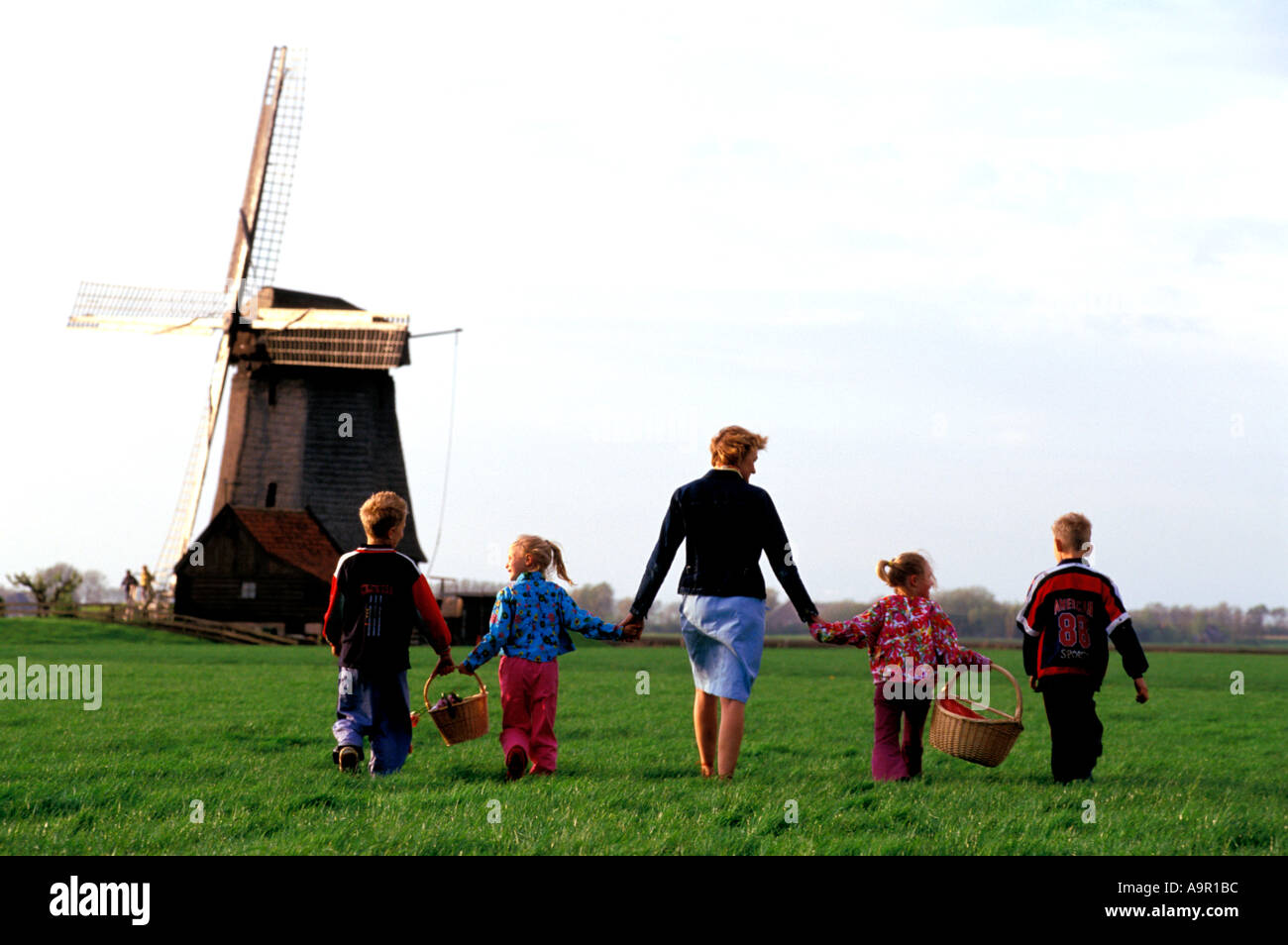 Mother and children with baskets crossing farmlands holding hands in Holland near windmill Stock Photo