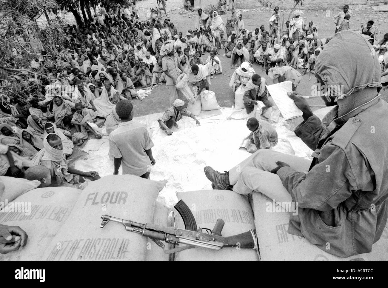B/W of a TPLF soldier with kalashnikov overseeing the distribution of food aid surrounded by people seated on the ground. Mekelle, Tigray, Ethiopia Stock Photo