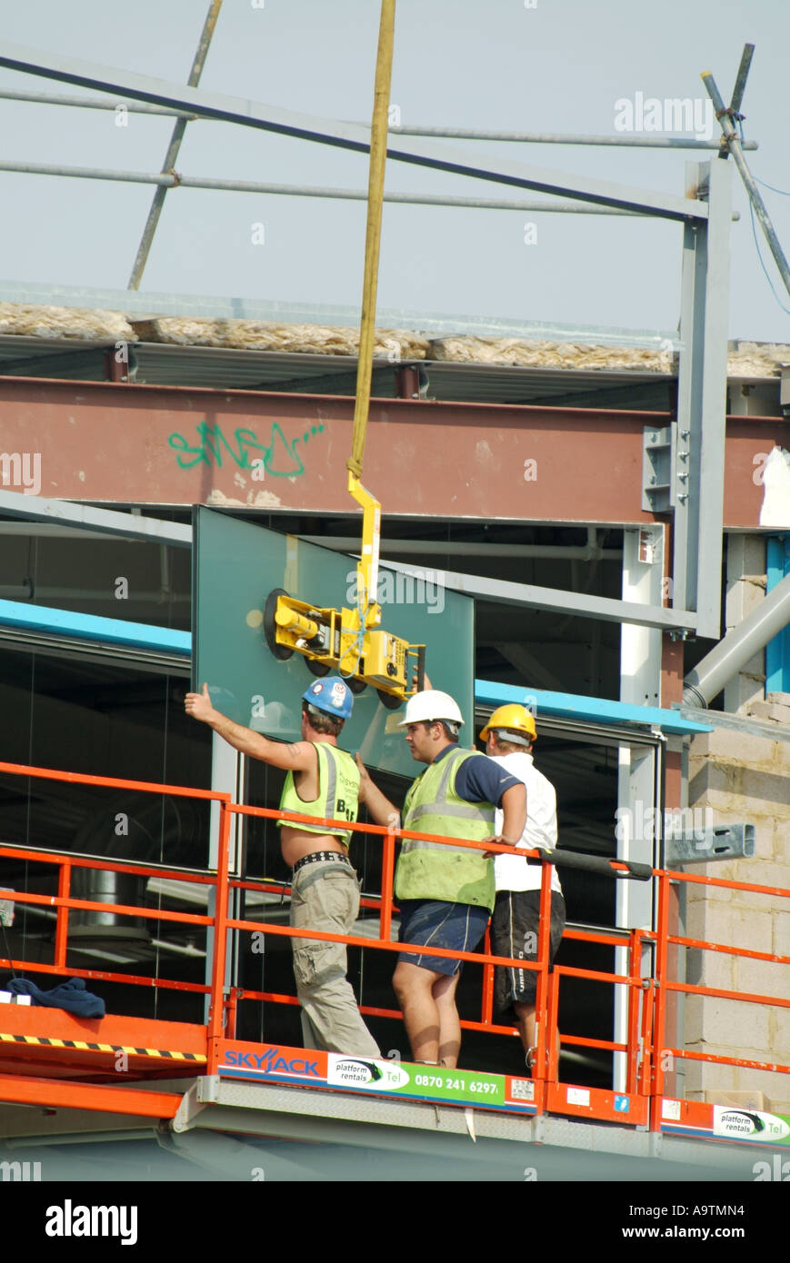 Workmen manoeuvring into position heavy cladding panel suspended from crane on warehouse construction site Stock Photo