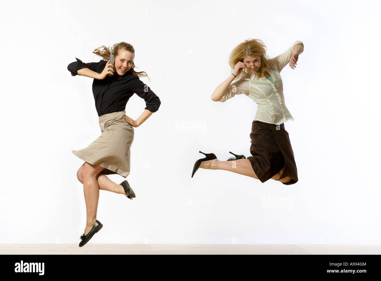 Two attractive young women dressed conservatively in business clothes jumping in the air. One is using a mobile phone/glasses Stock Photo