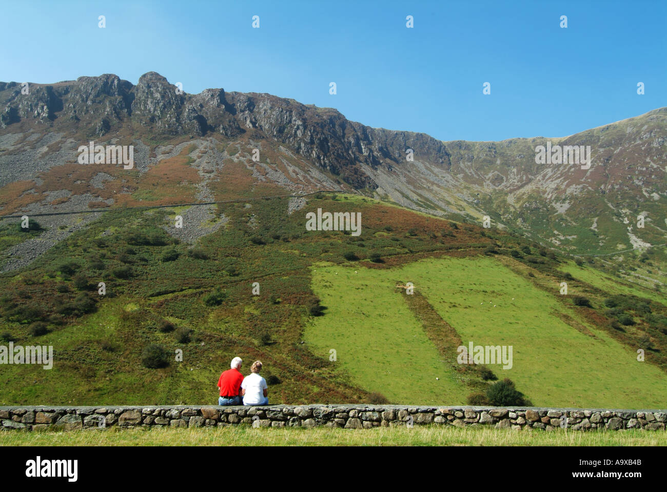Back view man & woman tourist couple sitting on stone wall in lay by view towards Cader Idris landscape on A487 road Snowdonia Gwynedd North Wales UK Stock Photo