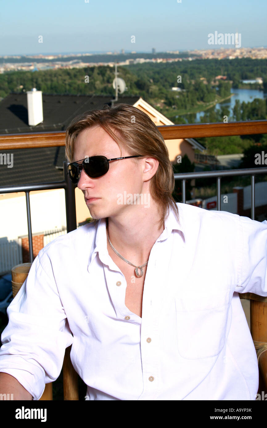 Stylish young man wearing sunglasses and a white shirt, sitting on a balcony with a scenic city and greenery view in the background. Stock Photo