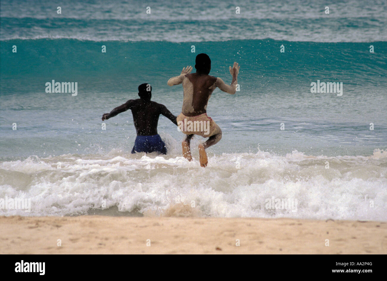 Two men jumping over surging waves and surf or wading into sea, Barbados, West Indies Stock Photo