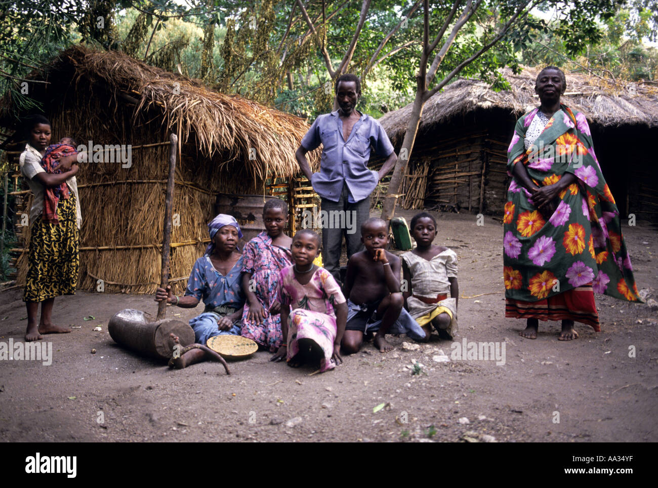 Extended family standing outside wooden huts, mother grinding rice. Uganda Africa Stock Photo