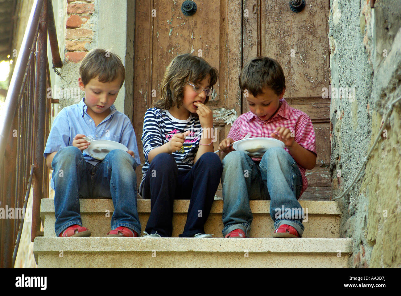 Three children eat cake from plastic plates on steps in rural Italy Stock Photo