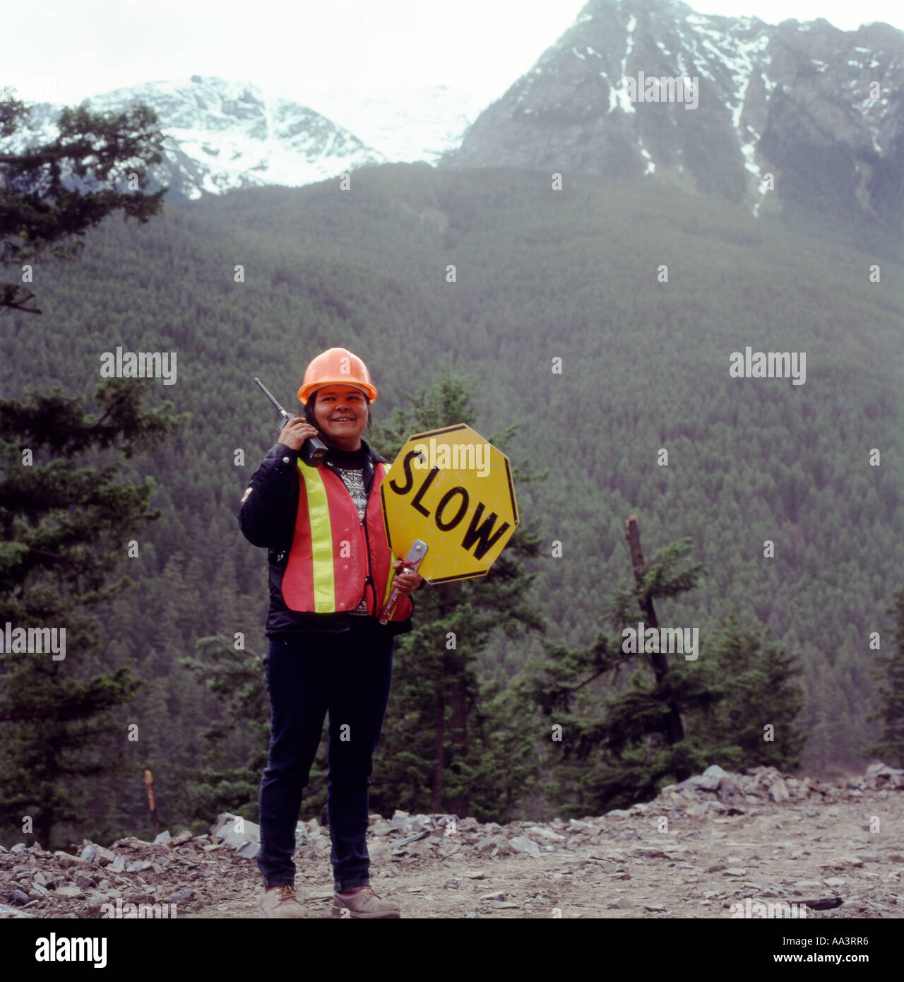 A First Nations female traffic worker woman holding a Slow traffic sign and cell phone on a forest road construction site in British Columbia Canada Stock Photo