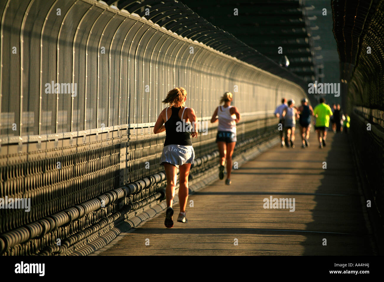 Joggers and runners run in the harsh industrial environment of the Sydney harbour bridge in the early warm morning light Stock Photo
