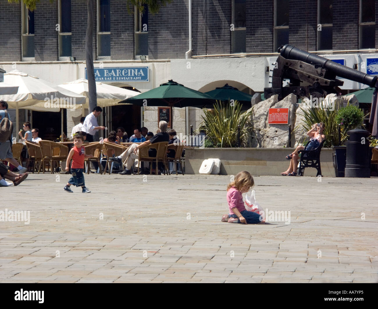 Little girl kneeling on the ground and boys playing football, Casemates Square, Gibraltar Stock Photo