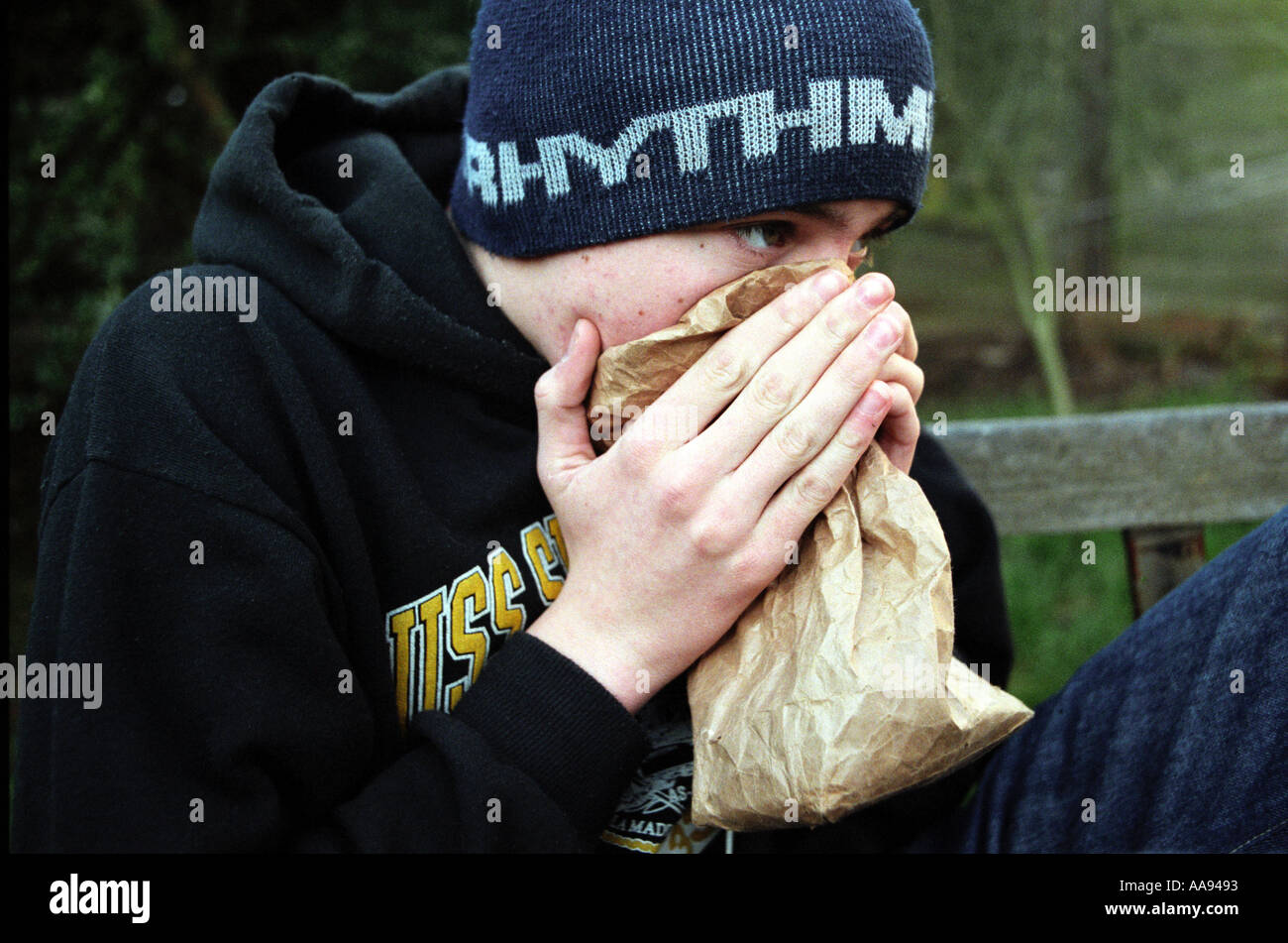 Young boy sitting on park bench sniffing glue from paper bag Stock