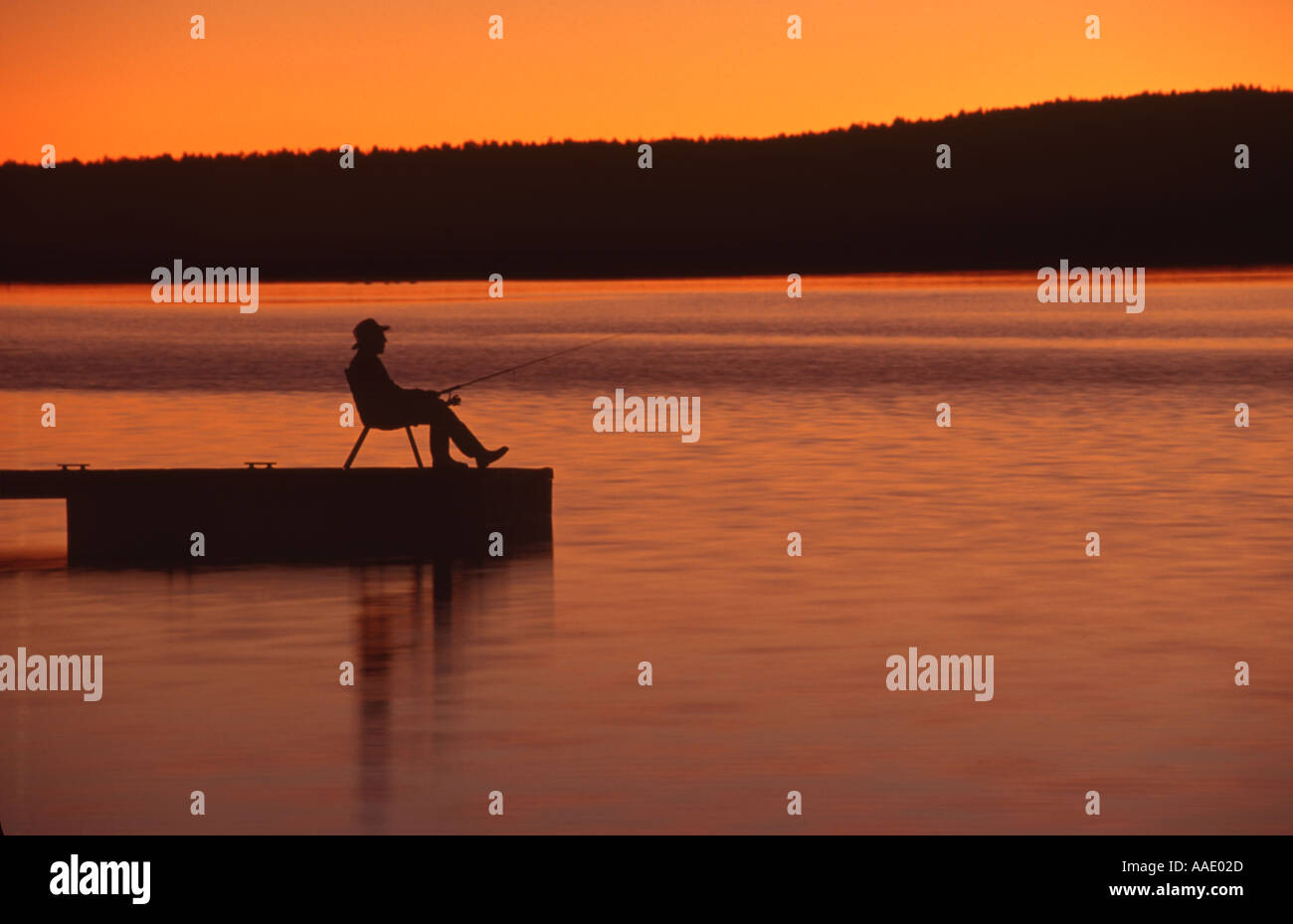 A fisherman fishes from a lake dock as he enjoys the sunset or sunrise Stock Photo