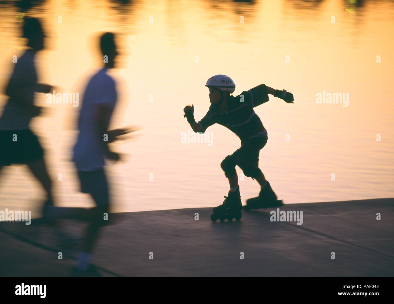 Joggers pass by a rollerskater along the shoreline of a city park at dusk Stock Photo