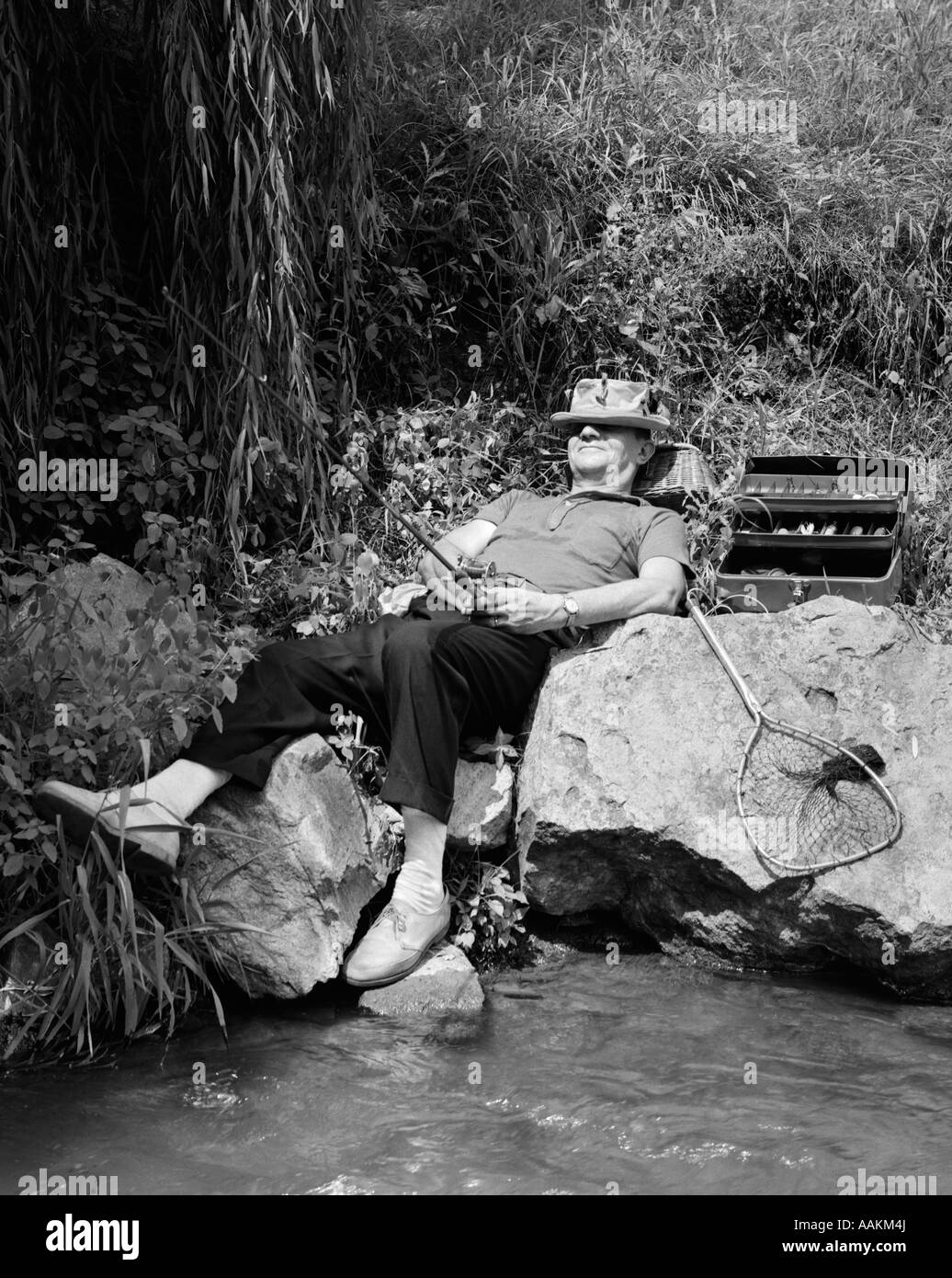 1950s LAZY FISHERMAN LYING BACK ON ROCK WITH HAT PULLED OVER EYES FISHING IN CREEK Stock Photo