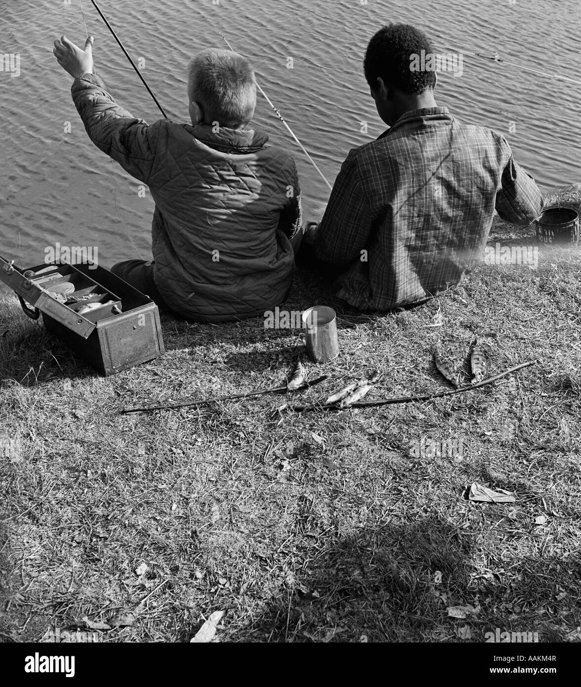 1960s BACK VIEW OF BLACK BOY AND WHITE BOY FISHING IN WATER WITH CAN OF WORMS & TACKLE BOX SHARED TOGETHER Stock Photo