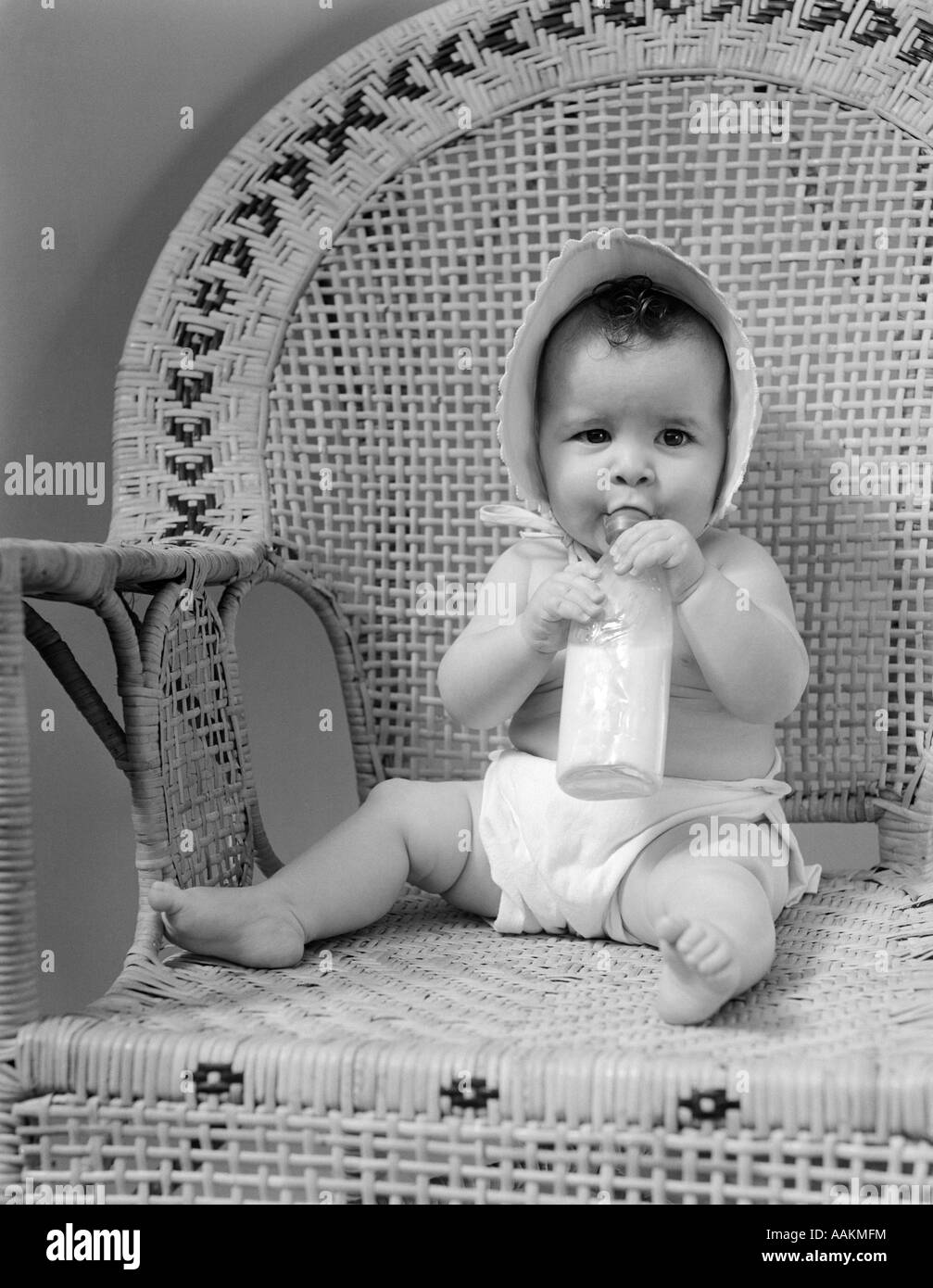 1940s BABY SITTING IN WICKER CHAIR SUCKING MILK BOTTLE Stock Photo