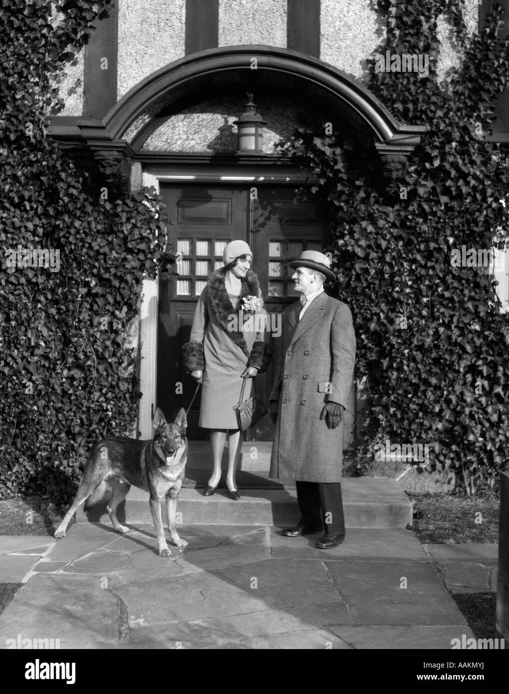 1920s COUPLE WEARING COAT HAT GLOVES ON STEPS IVY COVERED BUILDING WITH GERMAN SHEPHERD DOG ON A LEASH Stock Photo