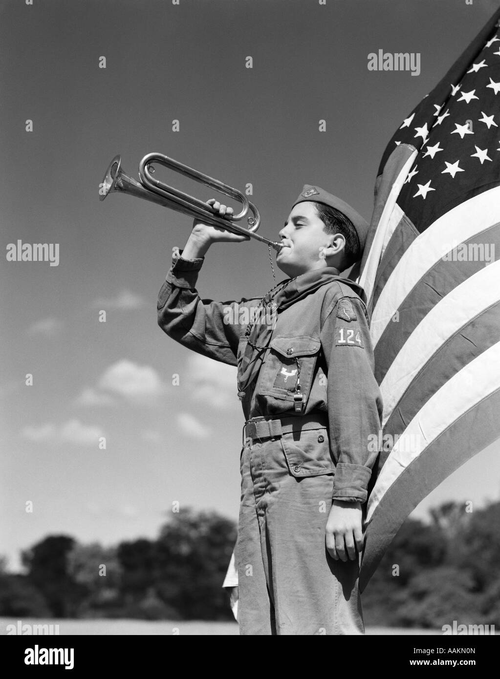 1950s PROFILE OF BOY SCOUT IN UNIFORM STANDING IN FRONT OF 48 STAR AMERICAN FLAG BLOWING BUGLE Stock Photo