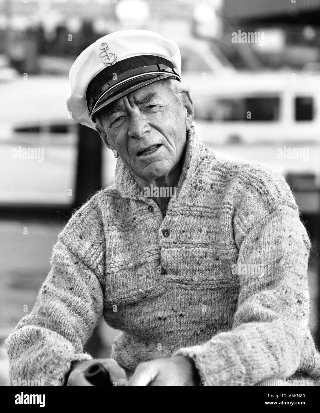 1970s OLDER MAN IN FISHERMAN'S HAT SWEATER HOLDING PIPE SITTING ON DOCKS OUTDOOR Stock Photo