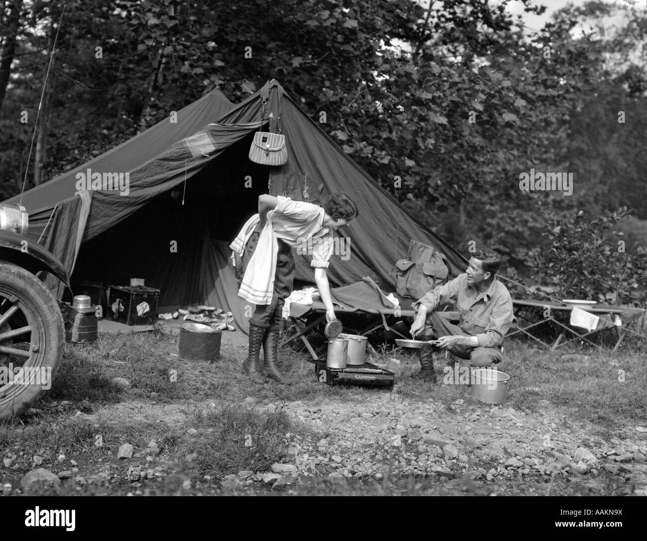 1920s COUPLE MAN WOMAN ON FISHING CAMPING TRIP VACATION AT CAMPSITE MAN COOKING OVER FIRE Stock Photo