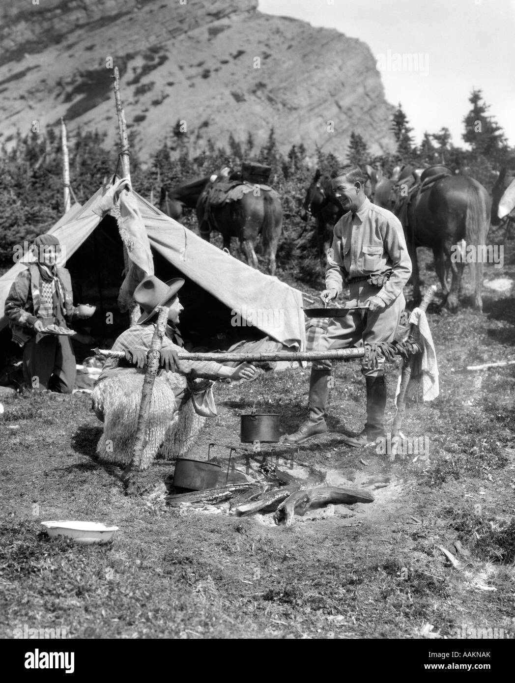 1920s 1930s TWO MEN AND ONE WOMAN EATING A MEAL AROUND A CAMPFIRE WITH A TENT AND HORSES AT BAKER LAKE ALBERTA CANADA Stock Photo