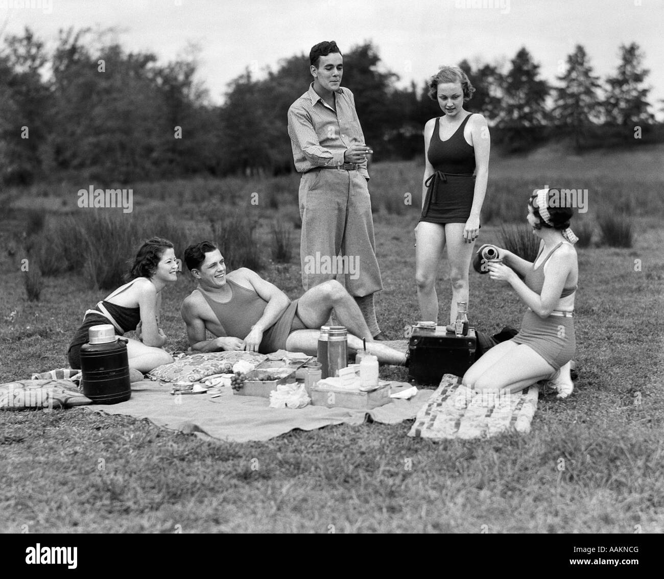 1930s GROUP OF FIVE YOUNG MEN & WOMEN ENJOYING PICNIC IN WOODS ALL BUT ONE WEARING SWIMSUITS Stock Photo