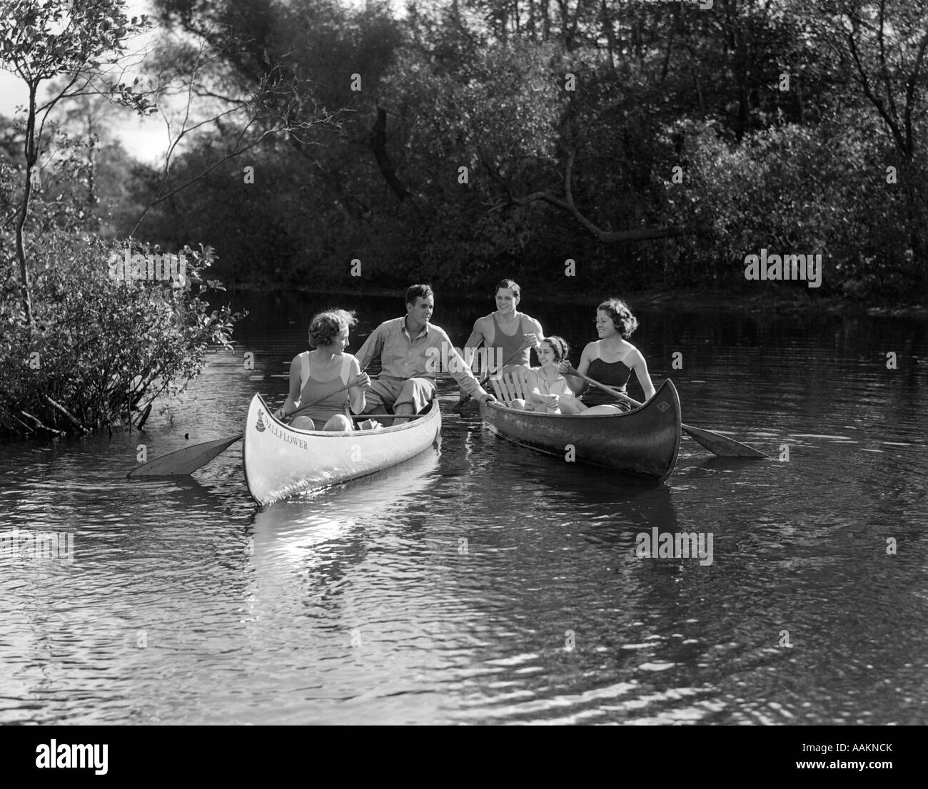 1930s SUMMERTIME GROUP OF FIVE YOUNG MEN & WOMEN IN TWO CANOES PADDLING DOWN A STREAM Stock Photo