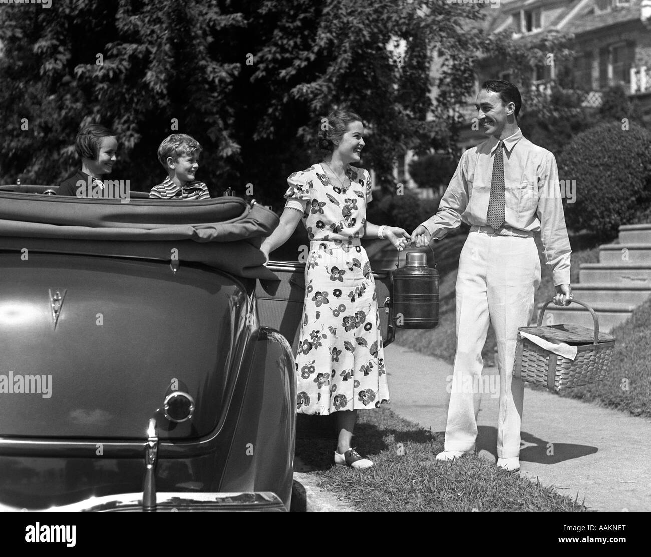 1930s FAMILY OF FOUR GETTING INTO CONVERTIBLE AUTOMOBILE WITH PICNIC BASKET AND THERMOS JUG Stock Photo