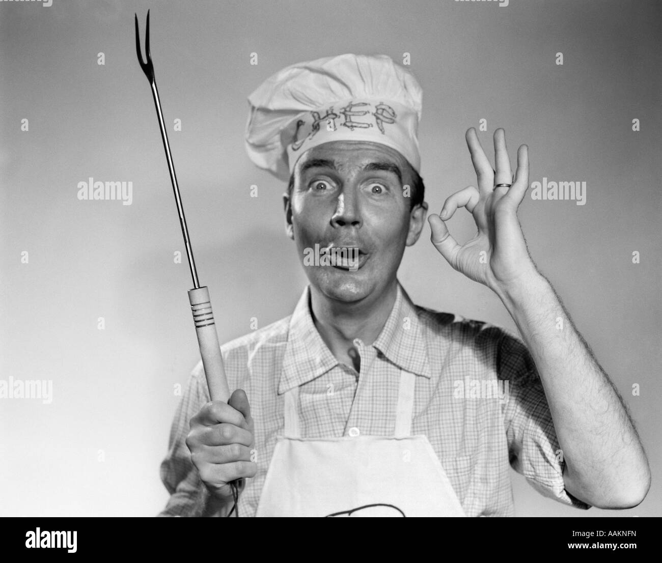1950s 1960s MAN WEARING CHEF HAT HOLDING BARBECUE GRILLING FORK MAKING OKAY SIGN LOOKING AT CAMERA Stock Photo
